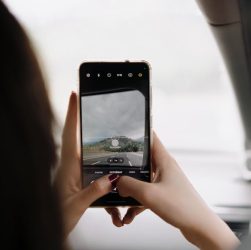 A person using a smartphone to capture a scenic view through the car window during a day trip.