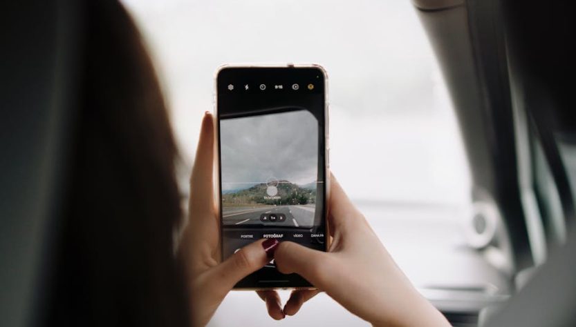 A person using a smartphone to capture a scenic view through the car window during a day trip.